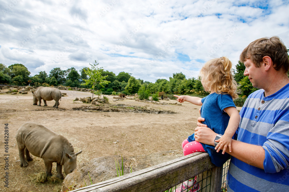 Fototapeta premium Cute adorable toddler girl and father watching wild rhinos in zoo. Happy baby child, daughter and dad, family having fun together with animals safari park on warm summer day. Ireland