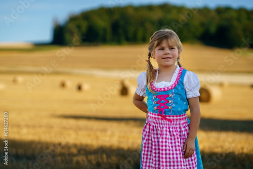 Cute little kid girl in traditional Bavarian costume in wheat field. Happy child with hay bale during Oktoberfest in Munich. Preschool girl play at hay bales during summer harvest time in Germany. © Irina Schmidt