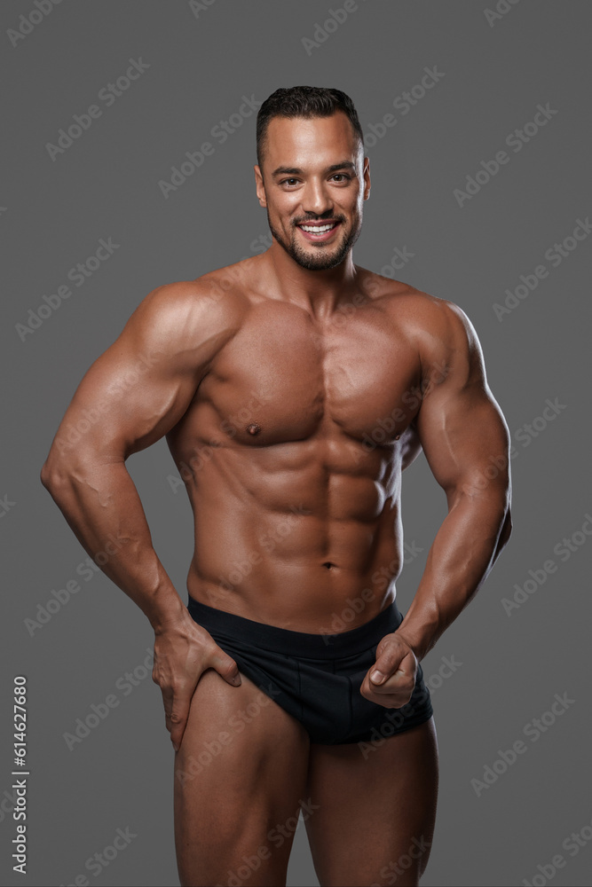 Breathtakingly handsome male model with a chiseled physique flashes a charming smile as he poses in black briefs against grey background, emanating charisma and sex appeal