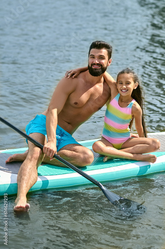 Handsome man with daughter sitting on surfboard in water enjoying vacation spending time together.