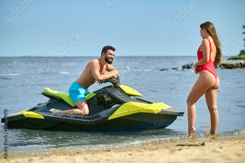 Couple communicating, man sitting on jet ski woman in red swimsuit standing on sandy shore.