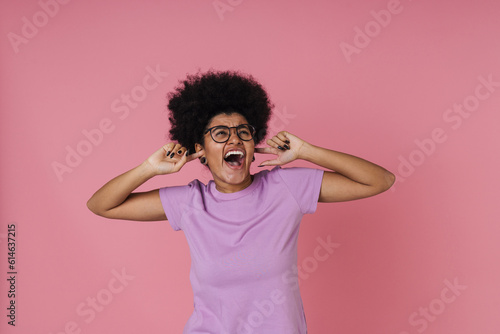 Annoyed woman covering her ears and screaming while standing isolated over pink wall