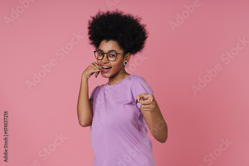 Cheerful african woman making call gesture and pointing finger at camera while standing isolated over pink wall