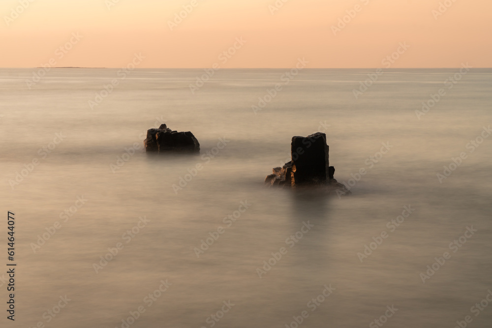 Photo exposes a marine landscape in an ancient lava rock rapids millions of years ago, at dusk, with two lava rocks protruding above the sea. Warm photo color. Soft focus.