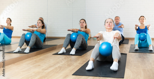Aged woman sitting on mat and doing sit-ups with bender ball during group training at gym. Healthy lifestyle and pilates concept