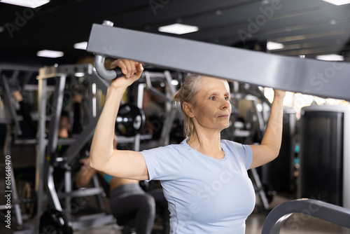 Concentrated fit elderly woman in sportswear working out in modern gym, performing exercises for back muscles building on lat pull down lever machine. Concept of healthy lifestyle of older generation
