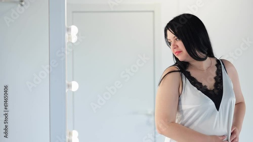 Black-haired chubby woman in t-shirt overweight lady looks at reflection on mirror at home. photo