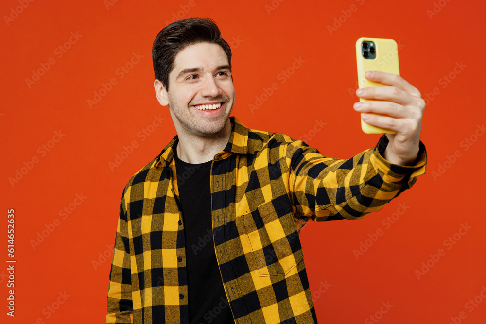 Young man he wear yellow checkered shirt black t-shirt doing selfie shot on mobile cell phone post photo on social network isolated on plain red orange background studio portrait. Lifestyle concept. Young man he wear yellow checkered shirt black t-shirt doing selfie shot on mobile cell phone post photo on social network isolated on plain red orange background studio portrait. Lifestyle concept.