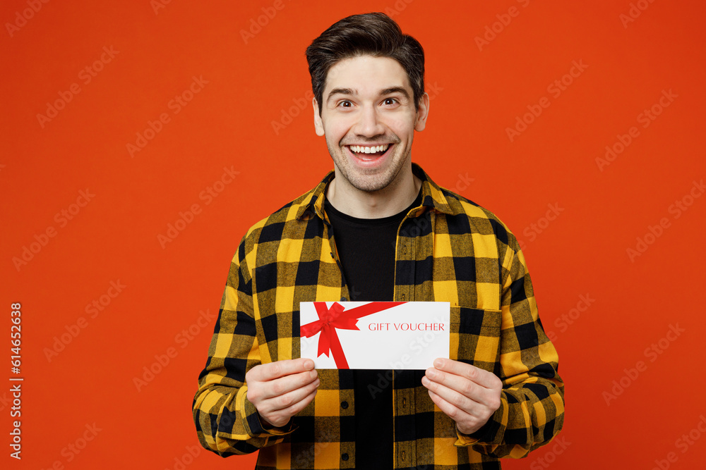 Young excited caucasian man he wear yellow checkered shirt black t-shirt hold gift certificate coupon voucher card for store isolated on plain red orange background studio portrait. Lifestyle concept. Young excited caucasian man he wear yellow checkered shirt black t-shirt hold gift certificate coupon voucher card for store isolated on plain red orange background studio portrait. Lifestyle concept.