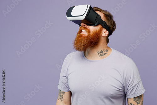 Young redhead bearded man he wear violet t-shirt casual clothes watching in vr headset pc gadget look aside on area isolated on plain pastel light purple background studio portrait. Lifestyle concept.