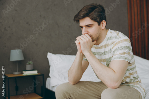 Side view young depressed frustrated disappointed man wear casual clothes t-shirt pajama sitting on bed propup chin think spend time in bedroom home in own room hotel wake up dream be in bad mood day. photo