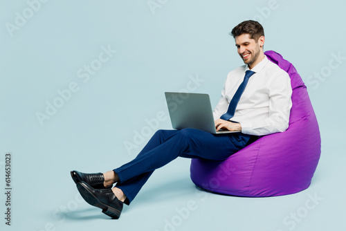Full body young employee IT business man corporate lawyer wears classic formal shirt tie work in office sit in bag chair hold use work on laptop pc computer isolated on plain pastel blue background.
