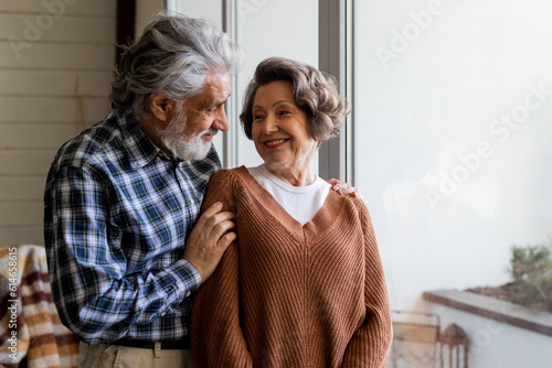 A couple of mature spouses cuddle nicely near a large window. An older man hugs his attractive mature wife.