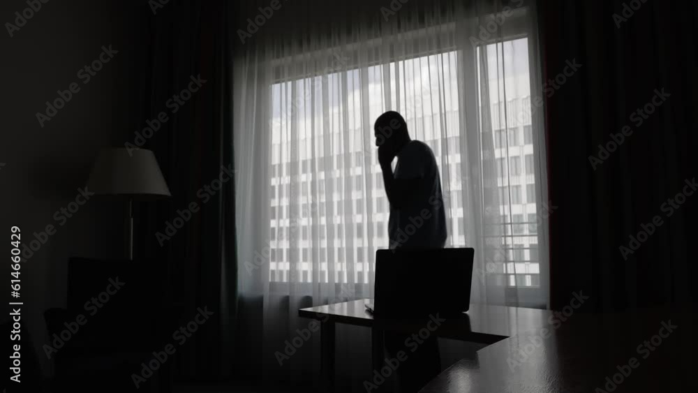 Silhouette Of Male Pacing Up And Down Beside Large Backlit Window In Hotel Room Talking Of Mobile And Gesturing At Laptop On Hotel Desk. Locked Off Silhouette Of Male Pacing Up And Down Beside Large Backlit Window In Hotel Room Talking Of Mobile And Gesturing At Laptop On Hotel Desk. Locked Off