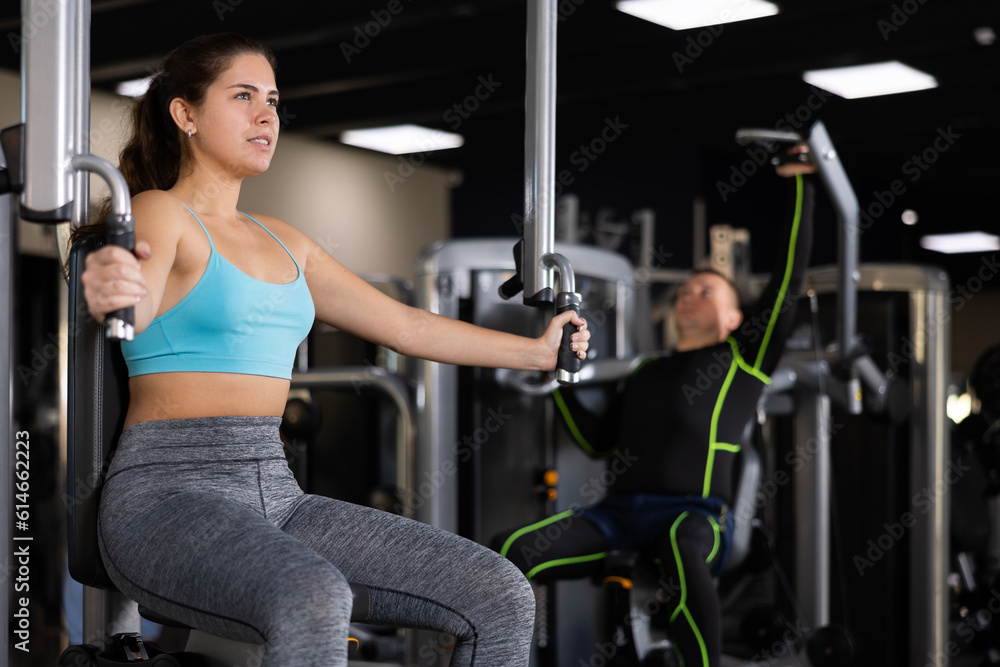 Fototapeta premium Concentrated young girl leading healthy active lifestyle doing strength training in gym, performing butterfly exercise for chest muscles in pec deck machine ..