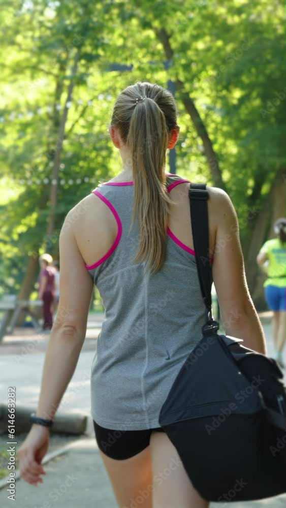 Vertical Screen: Woman with ponytail wearing sportswear carrying bag in public park, blurred people on background jogging, walking with dog and playing ping pong. Concept of healthy lifestyle Vertical Screen: Woman with ponytail wearing sportswear carrying bag in public park, blurred people on background jogging, walking with dog and playing ping pong. Concept of healthy lifestyle