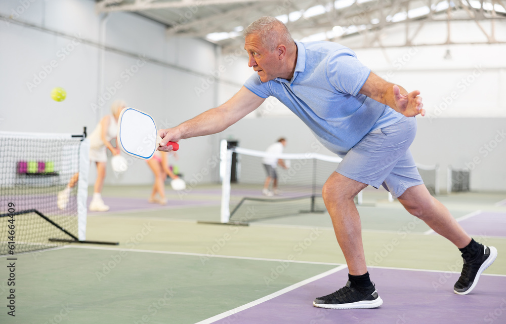 Portrait of active emotional elderly man playing pickleball on indoor court, swinging paddle to return ball over net ..