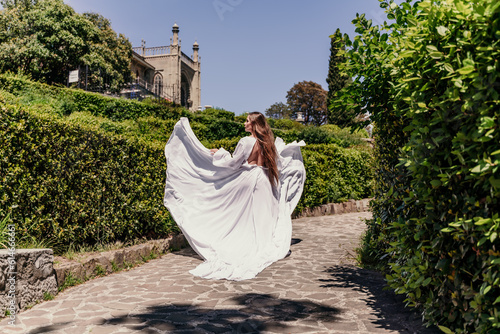 Brunette runs white dress park. A beautiful woman with long brown hair and a long white dress runs along the path along the beautiful bushes in the park  rear view