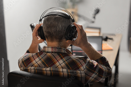 A man in a plaid shirt sits at a table in front of a laptop and a microphone. A man with gray hair puts on headphones and prepares to record a new podcast.
