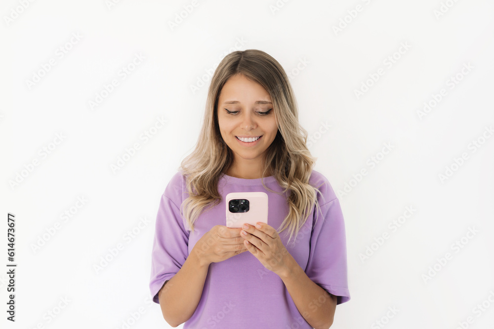 A young caucasian attractive happy pretty blonde woman in a purple t-shirt smiles and holds a mobile phone looking at the screen isolated on a white background. The joyful girl uses her smartphone A young caucasian attractive happy pretty blonde woman in a purple t-shirt smiles and holds a mobile phone looking at the screen isolated on a white background. The joyful girl uses her smartphone
