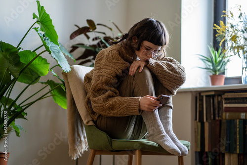 Upset teen girl sitting on chair with phone, hugging knees, scrolling on social media and feeling sad and unhappy. Depressed teenager with smartphone at home, gadgets and teenagers mental health