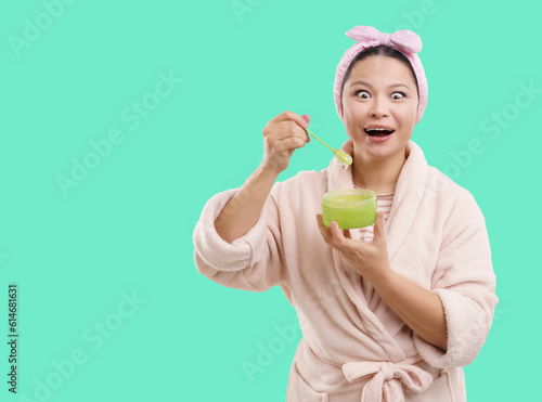 Excited Asian woman wearing bathrobe smiles at camera while opening cosmetic jar containing transparent cream. Isolated on vibrant green background, creating fresh and captivating visual. . High