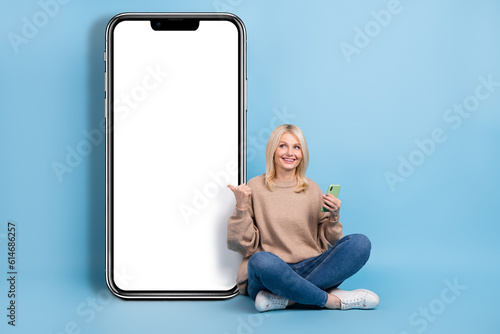 Full length body cadre of satisfied blonde hair sitting directing finger mockup smartphone board qr code isolated on blue color background