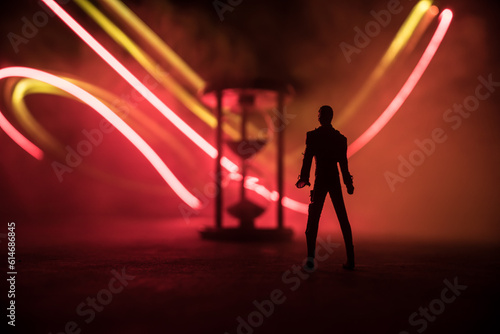Time concept. Silhouette of a man standing between hourglasses with smoke and lights on a dark background. © zef art
