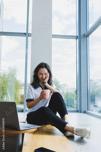 Free Download Smiling curly-haired woman sitting in a loft cafe, working at a computer, reading messages on a smartphone. Remote work - GFXLoad