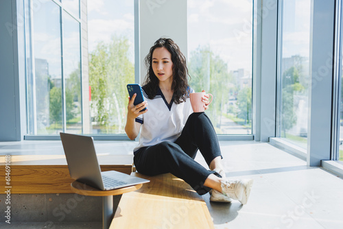 Free Download Smiling curly-haired woman sitting in a loft cafe, working at a computer, reading messages on a smartphone. Remote work - GFXLoad