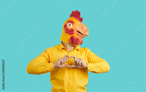 Fototapeta Naklejka Na Ścianę i Meble -  Funny man with comic romantic mood wearing chicken mask on his head shows heart shape showing love intentions. Eccentric man in orange shirt with absurd rubber mask chicken on light blue background.