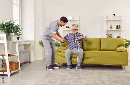 Portrait of a young nurse helping a senior patient with crutches to get up from the sofa at home. Physiotherapist helping old man in rehab. Health care worker helping an elderly man ta walk.