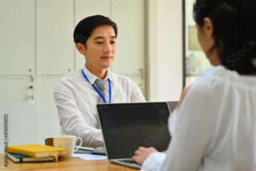 Image of asian man office worker using laptop computer in office with his colleague sitting on foreground 