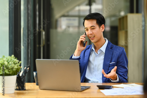 Young professional businessman wearing smart suit looking at laptop screen and talking on mobile phone in corporate office