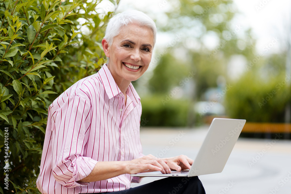 Portrait of successful senior businesswoman, freelancer using laptop computer sitting on bench outdoors. Attractive mature copywriter typing on keyboard working online at workplace. Business concept Portrait of successful senior businesswoman, freelancer using laptop computer sitting on bench outdoors. Attractive mature copywriter typing on keyboard working online at workplace. Business concept