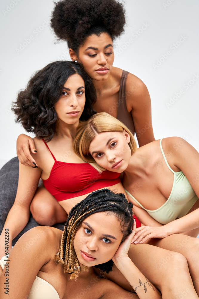 Fototapeta premium Multiethnic group of women with varied body shapes in colorful lingerie looking at camera and hugging while sitting on bed isolated on grey, different body types and self-acceptance concept