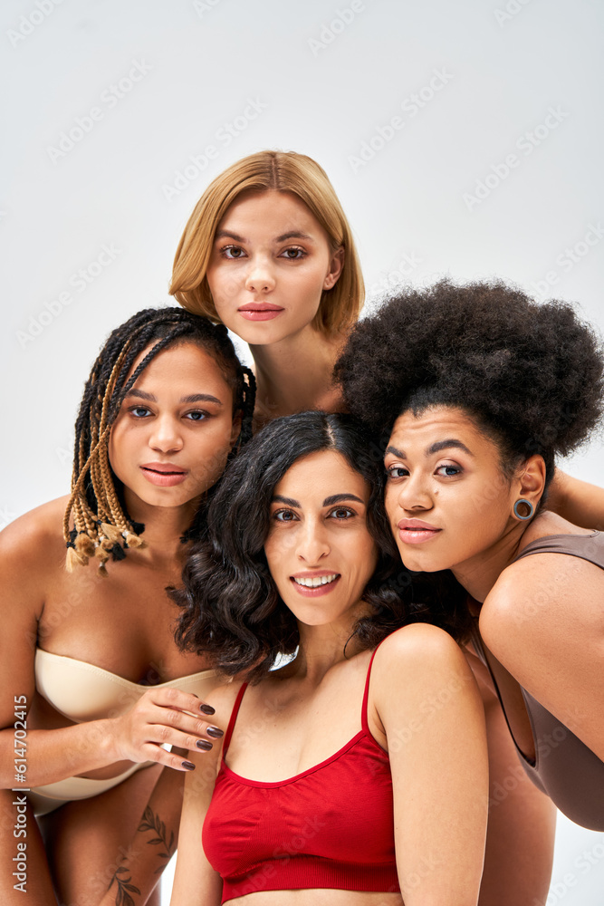 Positive multiethnic women in modern and colorful bras looking at camera together while posing isolated on grey, different body types and self-acceptance concept, multicultural models
