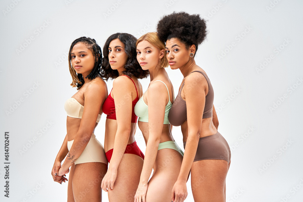Confident multiethnic women in colorful and modern lingerie looking at camera and standing together isolated on grey, different body types and self-acceptance concept, multicultural models