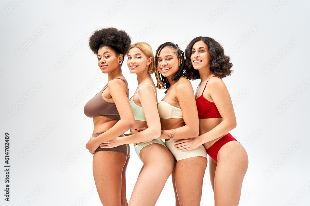 Joyful and multiethnic models in colorful and modern lingerie looking at camera and standing next to each other isolated on grey, different body types and self-acceptance concept, multicultural models