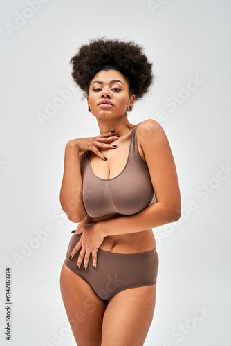 Portrait of seductive african american woman in modern brown lingerie touching neck and looking at camera while standing isolated on grey, self-acceptance and body positive concept