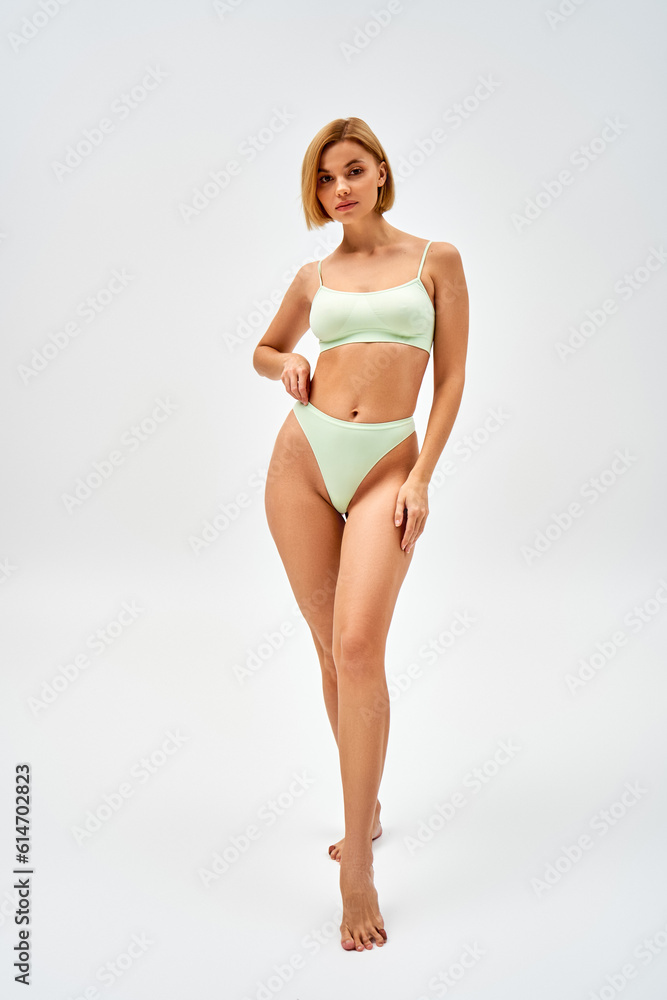 Full length of confident young blonde woman in light green lingerie looking at camera while standing and posing on grey background, self-acceptance and body positive concept Full length of confident young blonde woman in light green lingerie looking at camera while standing and posing on grey background, self-acceptance and body positive concept