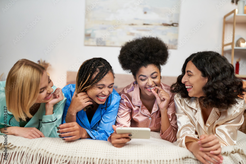 Laughing and multiethnic girlfriends in colorful pajama having fun while using smartphone together while lying on bed during pajama party at home, bonding time in comfortable sleepwear Laughing and multiethnic girlfriends in colorful pajama having fun while using smartphone together while lying on bed during pajama party at home, bonding time in comfortable sleepwear