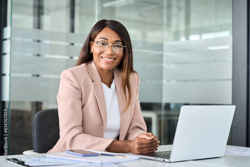 Young happy professional African American business woman wearing suit  eyeglasses working on laptop in office sitting at desk looking at camera,  female company manager executive portrait at workplace. Stock Photo