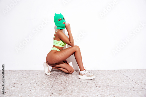 Beautiful sexy woman in green underwear. Model wearing bandit balaclava mask. Hot seductive female in nice lingerie posing near white wall in studio. Crime and violence. Sits on the floor