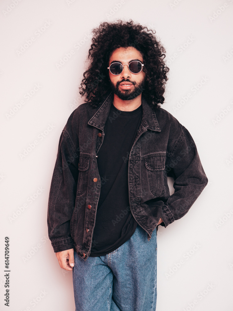 Handsome smiling hipster model. Sexy unshaven Arabian man dressed in summer stylish black jacket and blue jeans. Fashion male with long curly hairstyle posing near grey wall in studio. Sunglasses Handsome smiling hipster model. Sexy unshaven Arabian man dressed in summer stylish black jacket and blue jeans. Fashion male with long curly hairstyle posing near grey wall in studio. Sunglasses