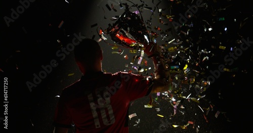 Silhouette of Caucasian male soccer football player raising a trophy above head against bright light and falling confetti. Super slow motion, shot on RED cinema camera