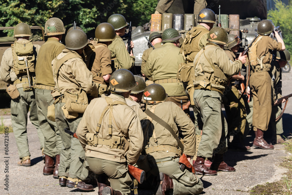Fototapeta premium Historical reconstruction. An American infantry soldiers from the World War II prepare to attack hidden behind a tank. View from the back.