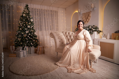 Owerweight elegant Woman at Christmas room. Fat plumb pretty girl in a beautiful dress for a holiday. Popular buxom female model posing alone in New year sudio