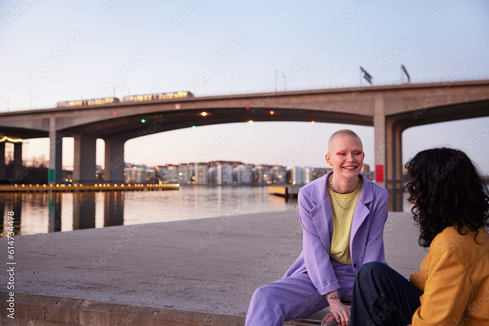 Young female gay couple sitting and talking at river, bridge in background Young female gay couple sitting and talking at river, bridge in background