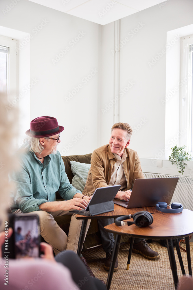 Senior men talking to each other while sitting in living room and using digital tablet and laptop to edit podcast Senior men talking to each other while sitting in living room and using digital tablet and laptop to edit podcast